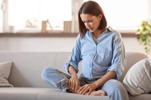 Young european pregnant lady in homewear massaging her foot indoor.